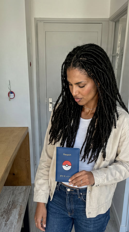 A woman with long black dreadlocks stands indoors holding a blue passport-style card featuring a red and white Pokéball logo. She is wearing a light beige jacket over a white top and blue jeans, with a gray door and wooden table visible in the background.
